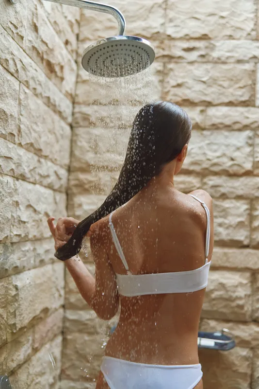 Woman showering outdoors under rain showerhead with long dark hair and white swimsuit, natural stone wall background, refreshing and relaxing moment.