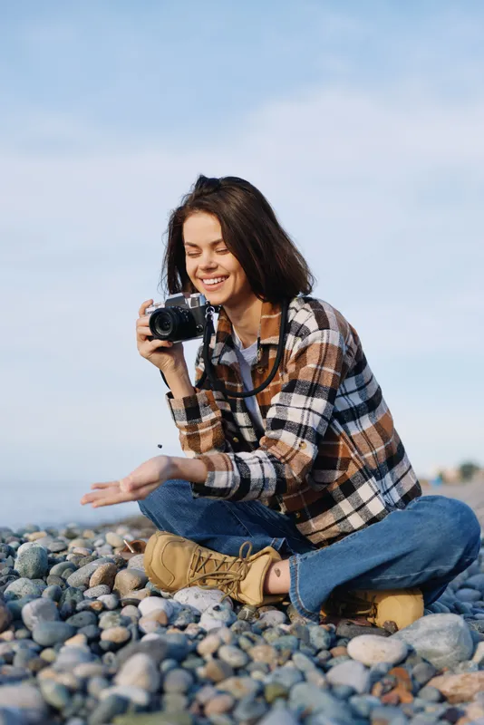 Photographer, camera and outdoors on a rocky beach, smiling woman in plaid shirt sits among pebbles capturing coastal moments with lens flare and relaxed travel vibe.