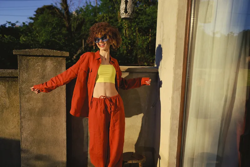A woman in orange tracksuit and lime top stands outdoors, radiating casual lifestyle energy under sunny light, with a modern balcony backdrop