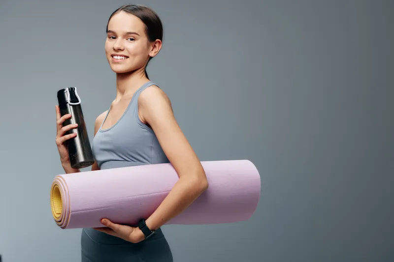 Woman in athletic wear holds a water bottle and a rolled yoga mat, ready for a workout. Fitness lifestyle photo featuring sport training, balance, and a focused, healthy routine.