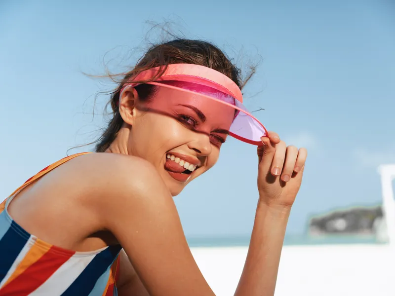Portrait of a joyful woman at the beach, wearing a colorful striped swimsuit and a pink headband, smiling under a clear blue sky.