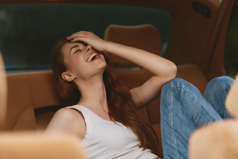 Joyful young woman laughing in a car, wearing casual jeans and a tank top, conveying happiness and relaxation in a cozy setting.