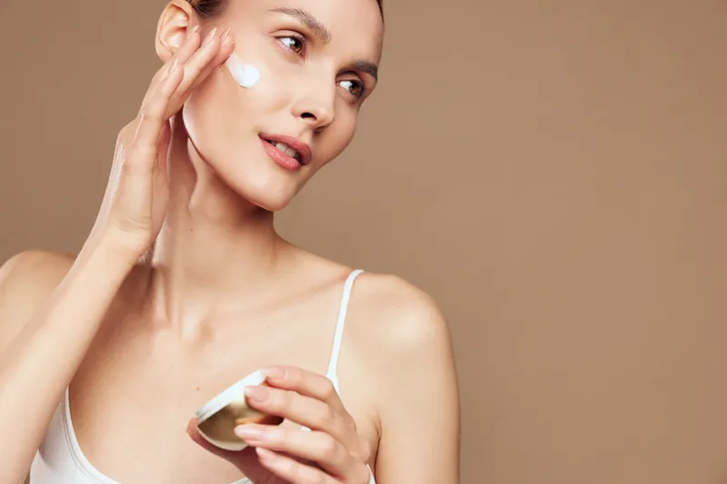 Young woman applies moisturizing cream on her face holding cosmetic jar with natural skincare product isolated on brown background with soft light and healthy skin concept.