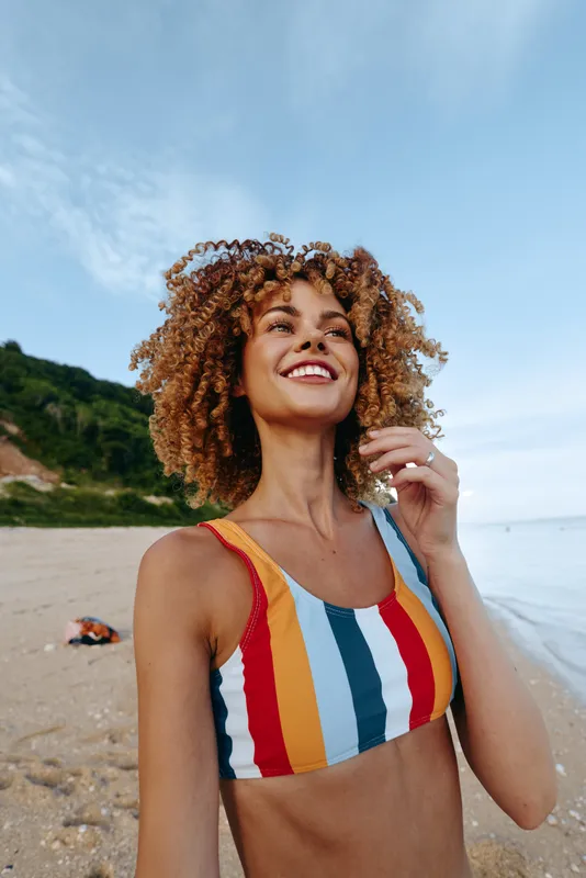 Woman with curly hair smiles on a sunny beach, wearing a multicolor striped swimsuit, standing by the sea, holding a delicate necklace, summer vacation travel, relaxed portrait outdoors