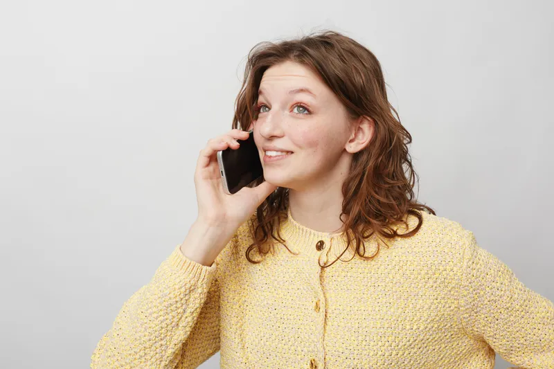 Young woman with curly hair wearing yellow sweater talks on smartphone smiling. Isolated on white background. Concept of communication, technology, casual lifestyle and positive mood.