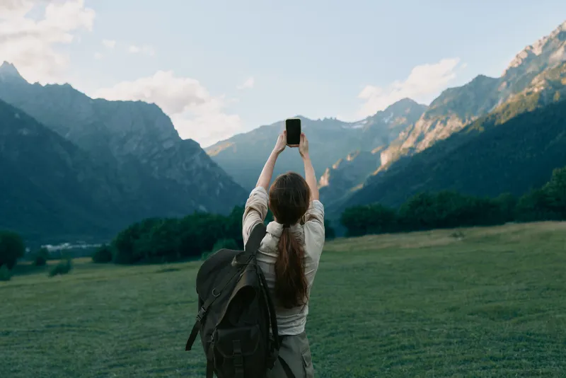 A woman with a backpack photographs a mountain valley in lush grass, capturing a solo travel moment. A lifestyle scene of exploration, outdoor photography, and freedom.