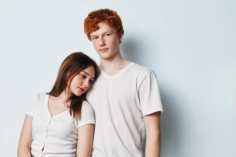 Young couple standing close together on white background. Woman leaning head on man shoulder with casual white t shirts. Friendship, relationship and affection concept with studio portrait.