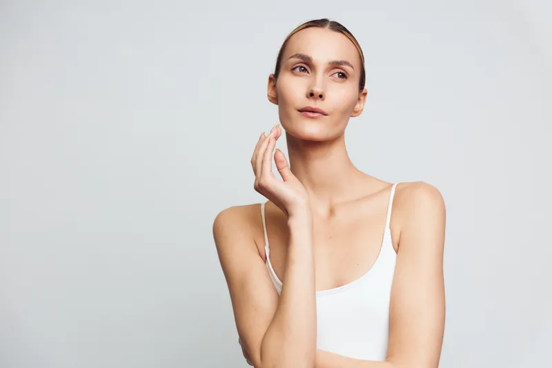Portrait of a thoughtful woman with natural skin wearing white tank top, posing against light gray background, minimal makeup, soft lighting, calm expression, clean aesthetic