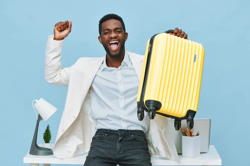 Smiling happy young black man in white blazer holding yellow suitcase look camera indoor on plain blue background studio portrait. Travel adventure lifestyle concept