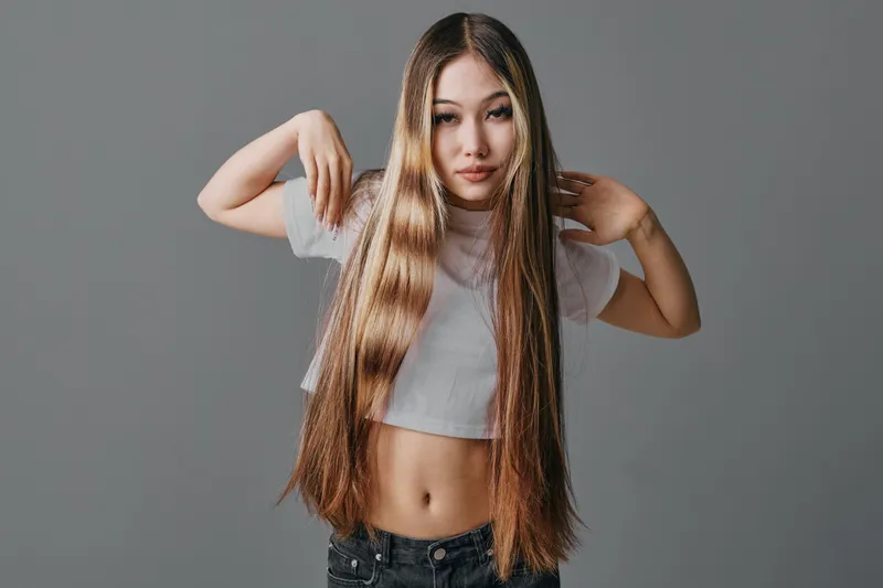 Girl with long hair, posing on gray background wearing a white crop top and dark pants. Attractive female model with straight hair showing casual confident expression and hand gestures.
