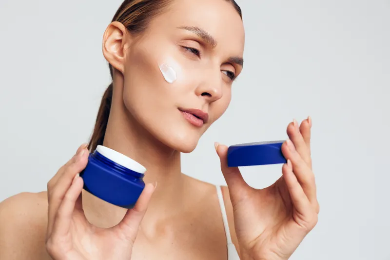 Young woman applies moisturizing cream on her face holding an open blue jar with white cosmetic cream isolated on white background with closed eyes and calm expression.