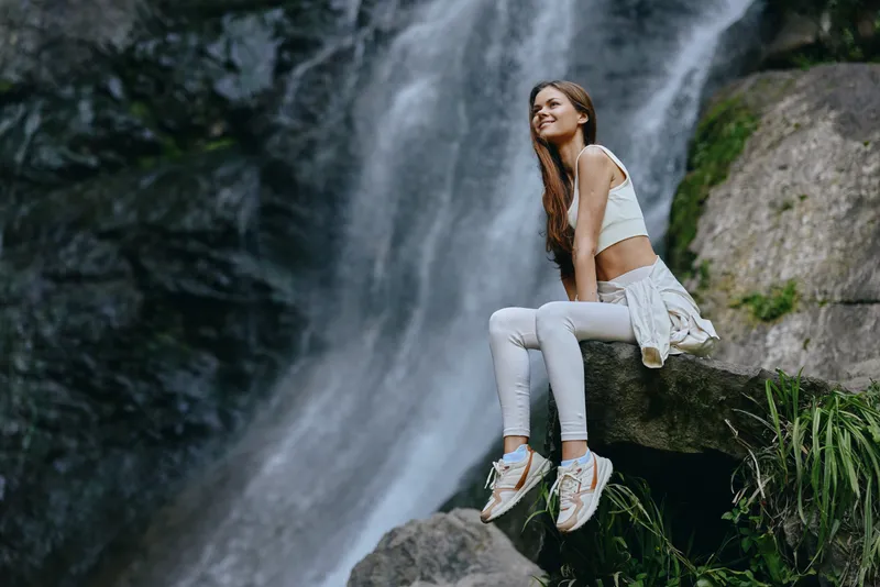 Smiling woman in white activewear sits on rock cliff near lush green plants with cascading waterfall in natural outdoor setting, radiating joy and serenity in soft daylight.