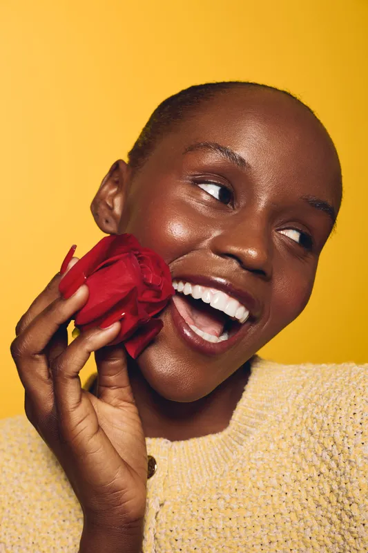 Smiling dark skinned African woman holds red rose near face with joyful expression on yellow background. Concept of beauty, happiness, natural skin, floral, and positive emotions.