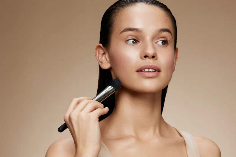 Young model applying makeup with a brush, showcasing natural beauty and elegance in a soft neutral background, perfect for beauty and fashion themes