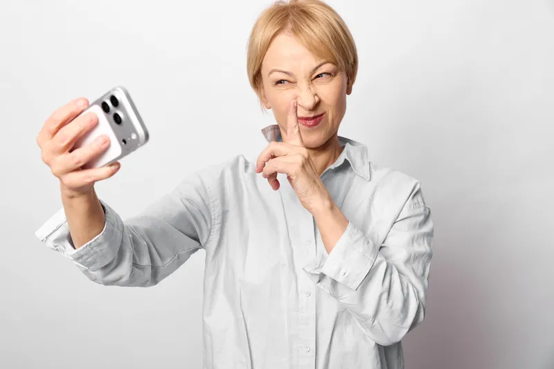 Elderly woman selfie with smartphone, smiling on a light gray background, portrait orientation. Gesture for communication and modern technology, friendly mood.