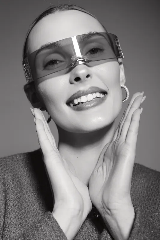 Smiling Caucasian woman with pale skin wearing futuristic sunglasses and earrings poses with hands near face in studio portrait. Fashion and style concept.