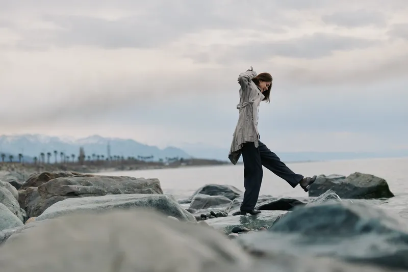 A young woman balances carefully on rocks by the sea under a cloudy sky. Casual outfit and natural coastal landscape create a peaceful and adventurous mood.