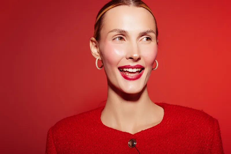 Smiling woman with red hair and red lipstick wears red sweater and hoop earrings. She looks away on red background. Concept of happiness, fashion, beauty, and vibrant style.