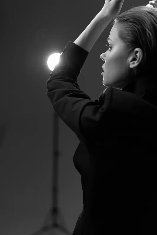 Black and white portrait of a thoughtful woman with her hands raised against studio light background in elegant outfit.