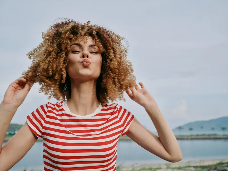 Playful portrait of a young woman with curly hair, wearing a red and white striped shirt, blowing a kiss toward the camera by a calm seaside backdrop.