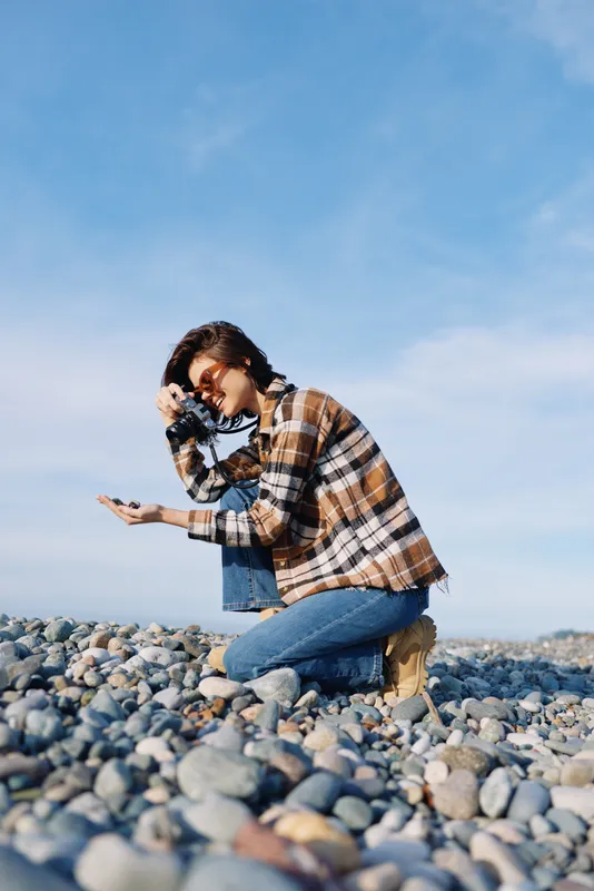 Photographer kneels on rocky beach, aiming a camera at the shoreline, wearing a plaid shirt and denim, capturing a candid outdoor portrait against a clear blue sky and light breeze
