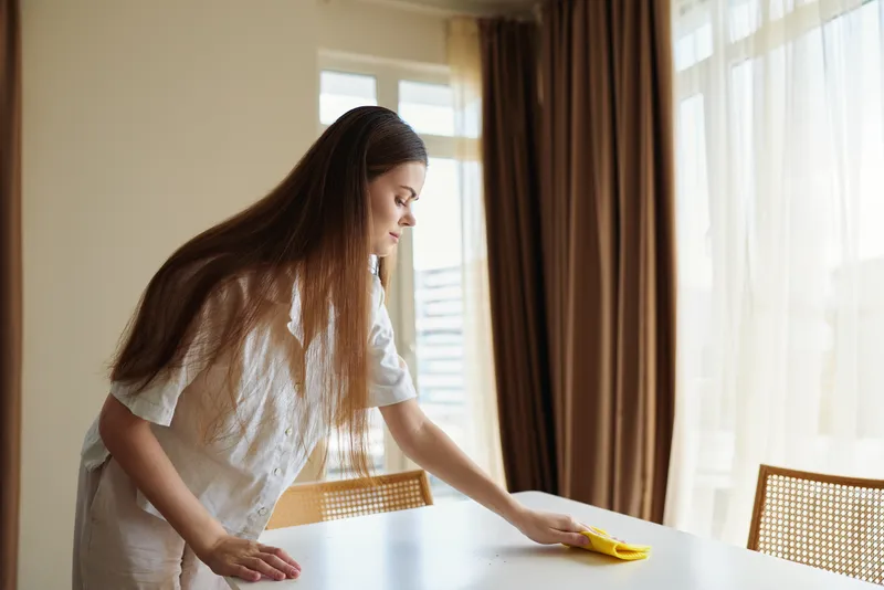 Woman cleaning table with yellow cloth in bright home interior by window and curtains, wiping surface for tidying and household chores, domestic routine and care.