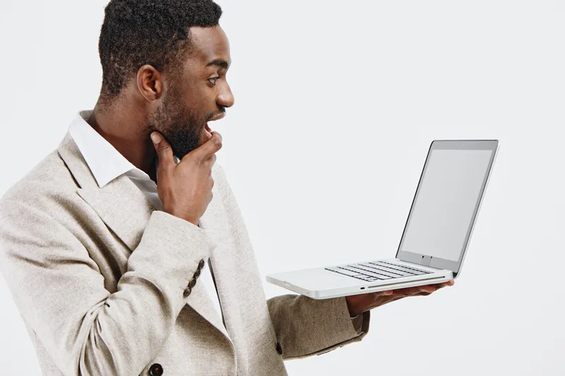 Thoughtful young African American man in beige blazer holding open laptop computer with blank screen, looking at it with curiosity in modern studio setting. Business technology concept for remote work