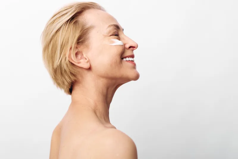 Smiling middle aged woman with short blonde hair applies moisturizing cream on cheek in bright studio. Concept of skincare, anti aging, self care and natural beauty for mature skin.
