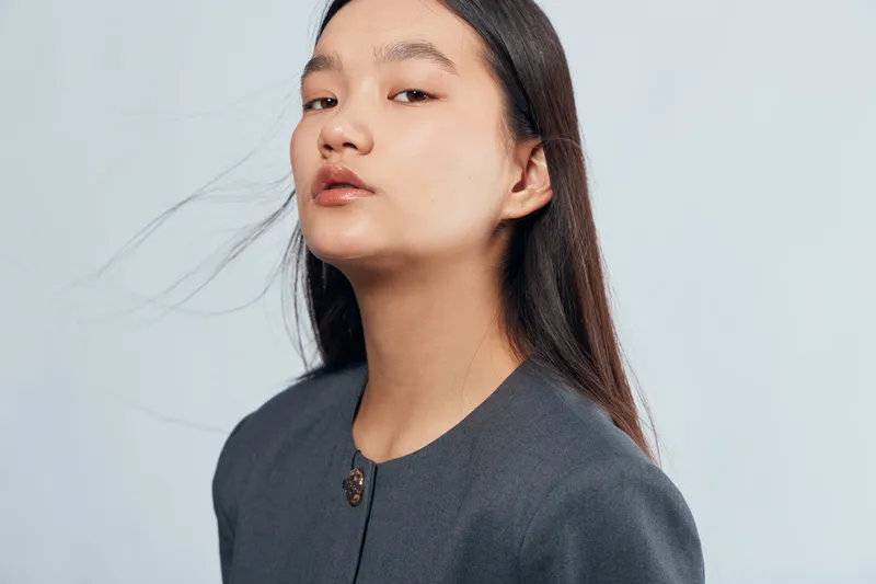 Portrait of confident asian woman with long dark hair wearing dark gray jacket standing on light gray background looking ahead with slight tilt of head and calm facial expression.
