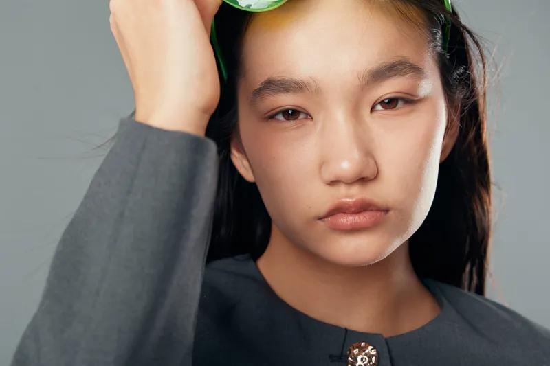 Young woman with natural beauty wearing gray outfit posing with serious expression and hand near sunglasses in studio portrait with plain background and soft lighting.