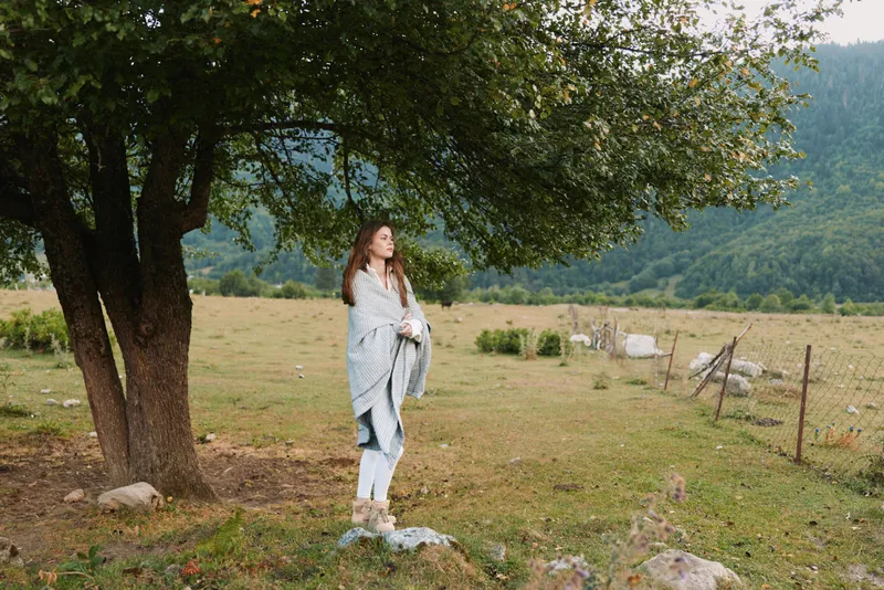 Cheerful woman outdoors, lifestyle moment in the countryside, standing under a large tree in a sunlit meadow, with a gentle breeze and natural colors