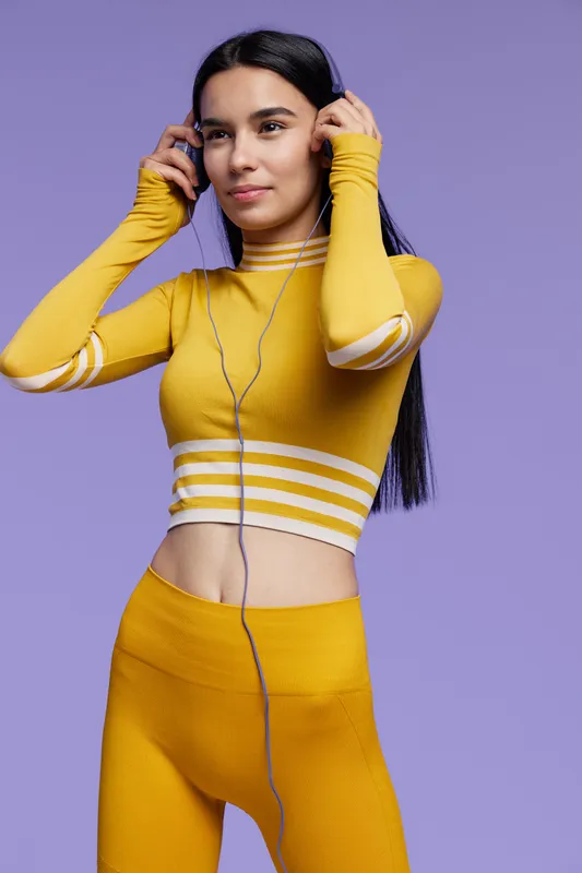 Fit woman sport fitness wearing bright yellow activewear adjusting headphones, preparing for workout or exercise session against a purple background.