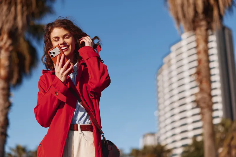 Young woman with curly hair smiles happily while holding a smartphone outdoors on a sunny day with palm trees and modern buildings in the background.