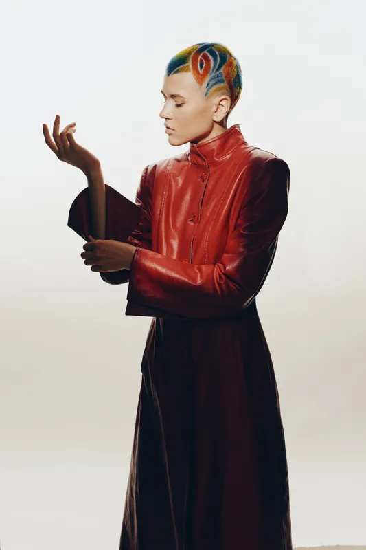 Woman with colorful shaved hair in red leather coat poses creatively against plain white background, showcasing bold fashion and artistic expression with serious mood.