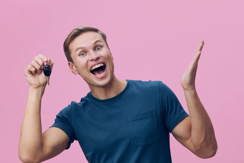 Joyful young man expressing excitement while holding car key on pink background. Casual blue t-shirt, cheerful facial expression, lifestyle concept, happiness, celebration, success, enthusiasm, positi