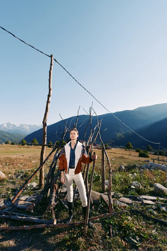 Man in traditional clothing stands by a rustic fence in a rural mountain landscape and grassy field under clear sky. Outdoor portrait, countryside lifestyle, scenic nature scene.