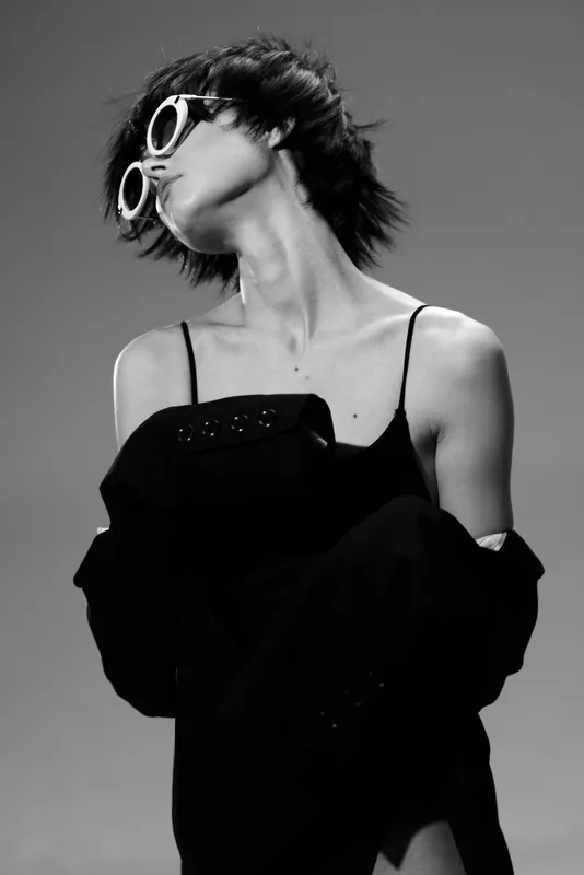 Black and white portrait of a young woman wearing sunglasses and stylish black dress with off-shoulder sleeves, showing confident attitude.