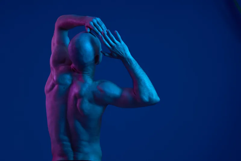 A striking image of a man posing dramatically in an artistic and colorful light, showcasing strong muscles and expressive body language in a unique blue and pink atmosphere.