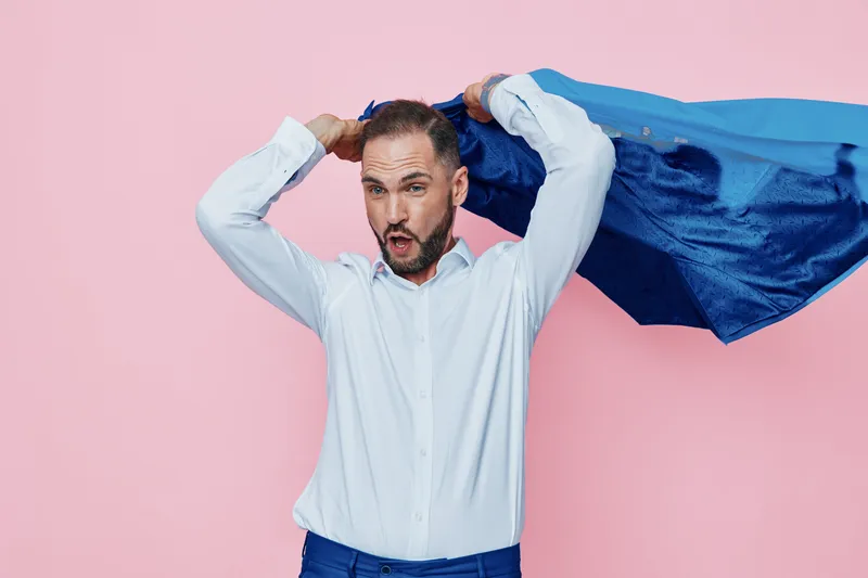 A business professional poses against a solid pink background, dressed in a white shirt and dark trousers, holding a blue cape as a playful flight gesture to express energy and pride.