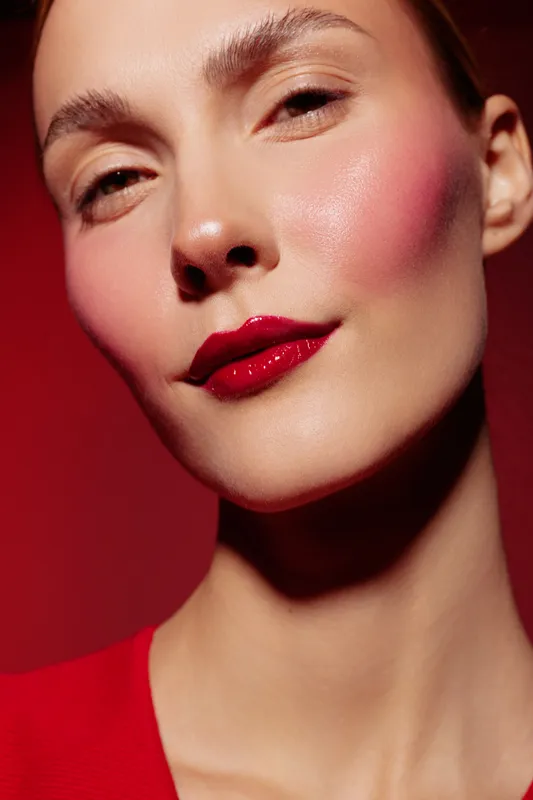 Close-up portrait of a woman with glossy red lips and rosy cheeks against a red background. Concept of beauty, makeup, skincare, and fashion with soft lighting and smooth skin.
