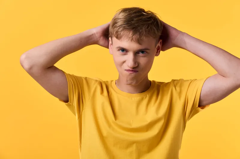 Confused frustrated puzzled expression of a young male with hands on head wearing a yellow shirt against a bright backdrop, conveying doubt, stress and hesitation in a simple studio shot