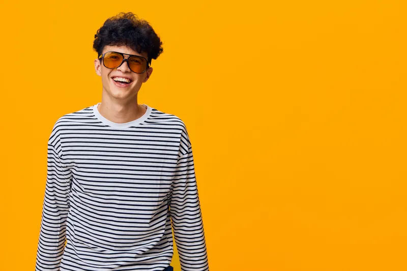 smiling young man wearing sunglasses and striped shirt laughing against bright orange background, casual style and joyful expression, studio portrait
