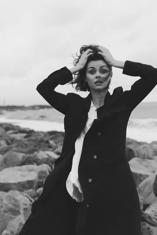 Fashion, coat, woman, wind, beach, ocean, blackandwhite, dramatic mood as waves crash near rocks, movement and runway attitude in a candid seaside moment