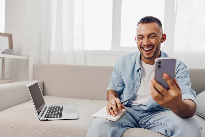 Smiling man engaged in a video call, expressing joy and enthusiasm. He is casually dressed and appears relaxed in a modern home setting.