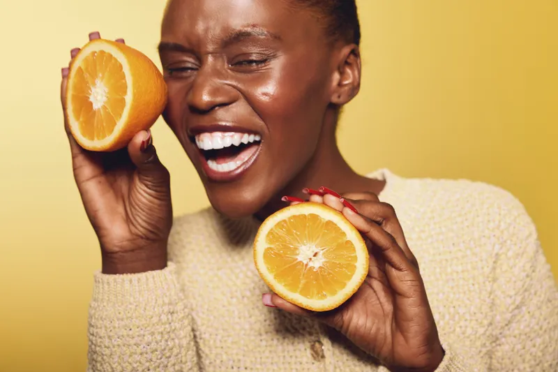 Joyful dark skinned African woman holds two orange halves near her face smiling with eyes closed. Concept for healthy lifestyle, fresh fruit, vitamin C and natural beauty on yellow background.