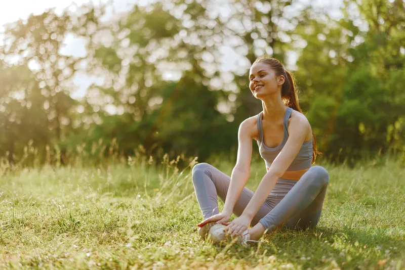 Young athletic woman stretching outdoors in a sunny park, embodying fitness and wellness with a joyful expression, perfect for healthy lifestyle promotion