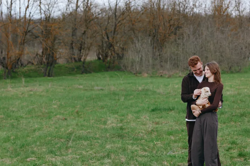 A couple smiles in a green field, holding a gentle goat kid, enjoying a casual countryside moment. A lifestyle portrait of warmth, family bond, rural charm, and peaceful outdoor living in daylight.