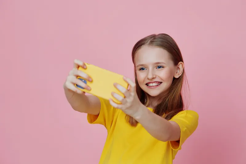 Happy young girl taking a selfie with a smartphone against a pink background, wearing a bright yellow t shirt, showcasing joy and vibrancy