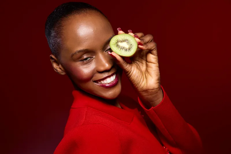 Smiling black woman with dark skin holds half of a kiwi fruit near eye on red background. Portrait of happy African female model in red sweater with fresh fruit concept of health and wellness.
