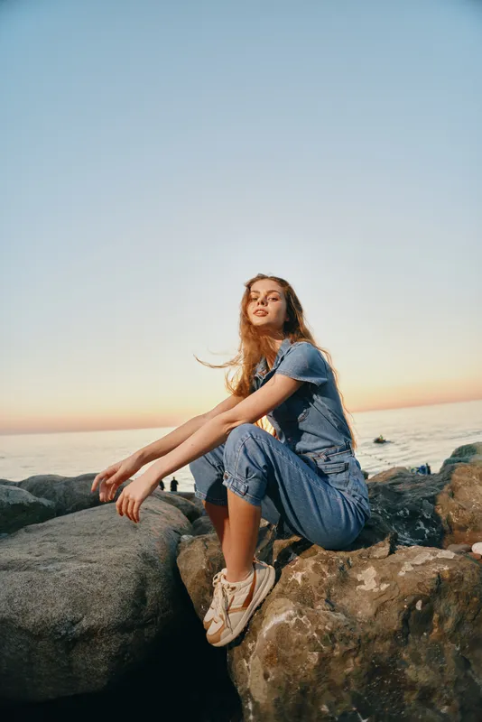 Lifestyle woman in modern boho-western denim street style sitting on rocks by the sea at sunset, captured with warm film color tones and casual relaxed pose.